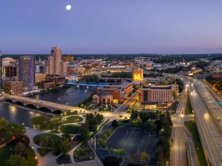 City skyline at dusk with a river, bridges, and illuminated buildings under a rising moon.