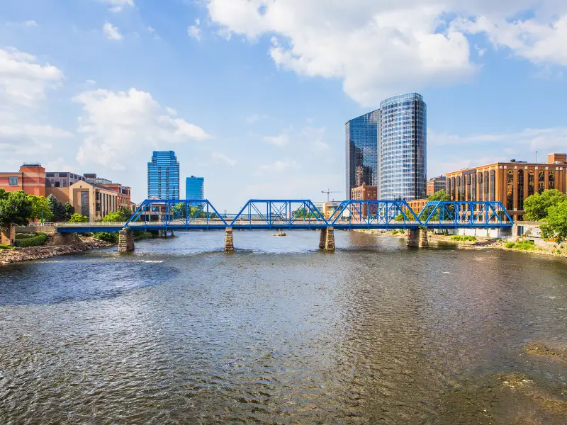 Blue steel bridge spanning a river with modern buildings and a partly cloudy sky in the background.