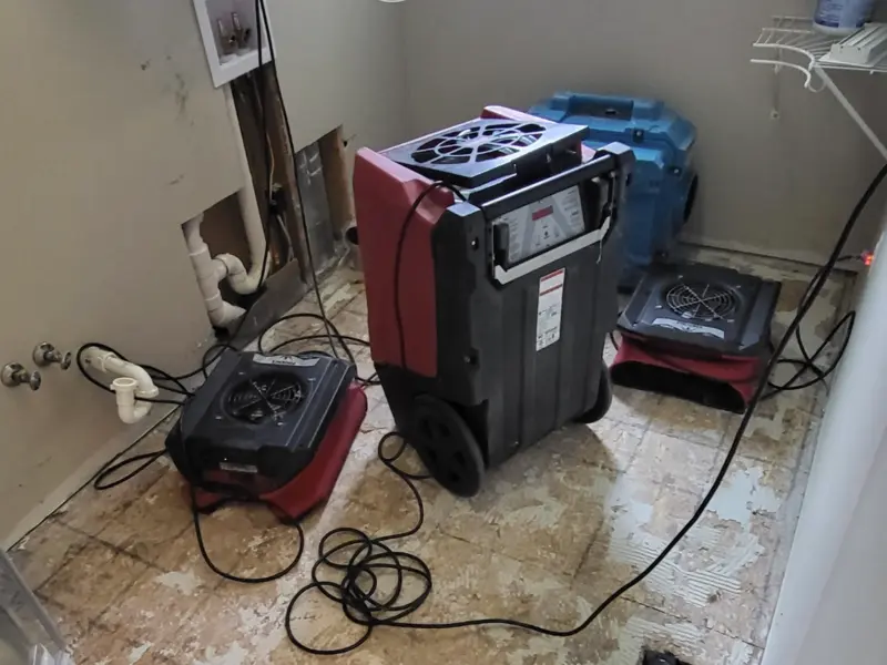 Water damage drying equipment set up in a room with exposed pipes and bare floor.