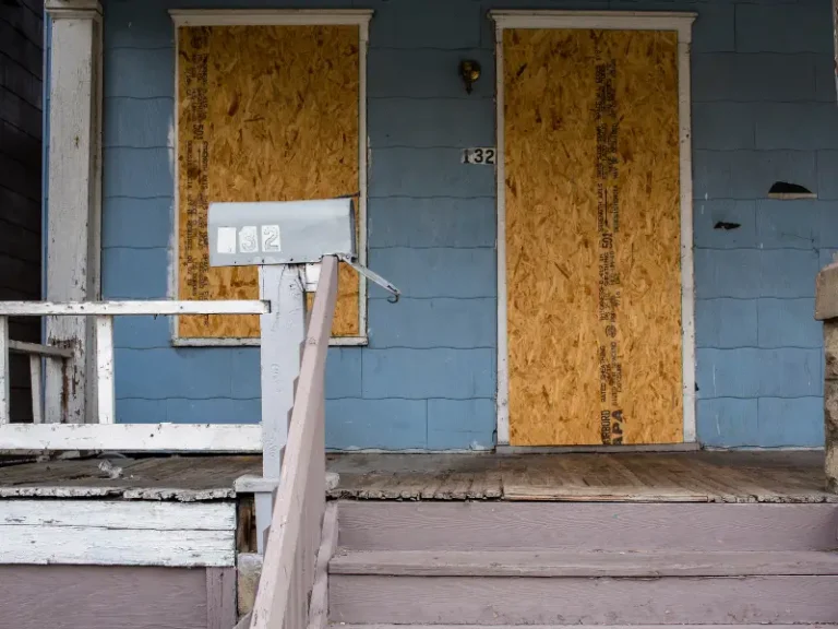 Boarded-up blue house with a mailbox and peeling paint on the porch steps and railing.