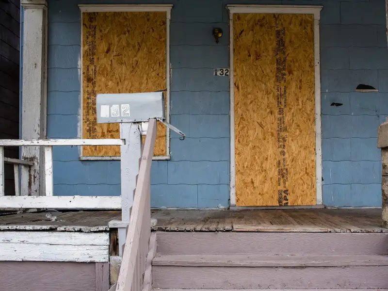 Boarded-up blue house with a mailbox and peeling paint on the porch steps and railing.