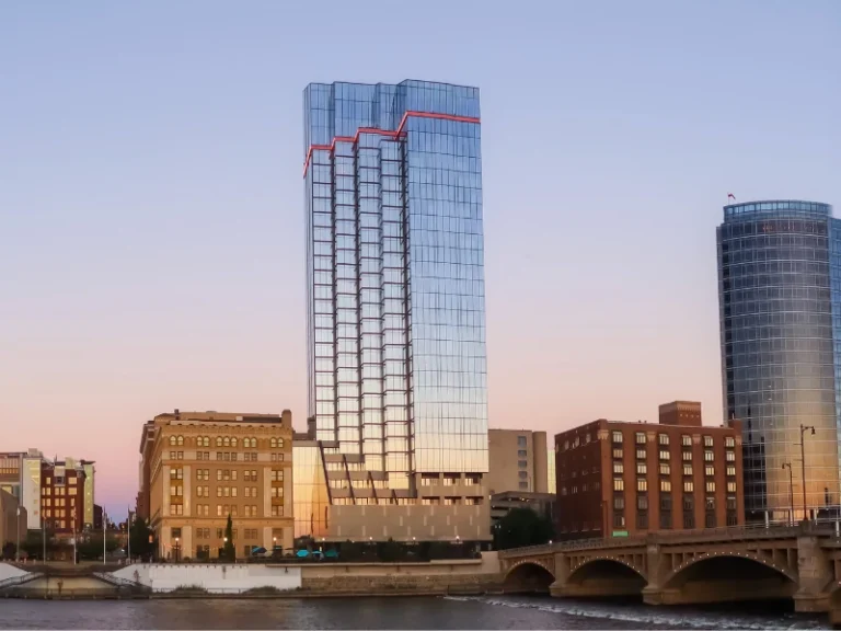 Glass skyscraper with red lights on top beside a river and a bridge at sunset.
