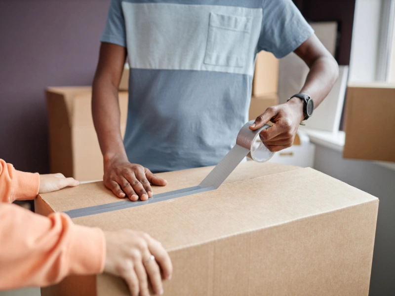 Two people sealing a cardboard box with packing tape.