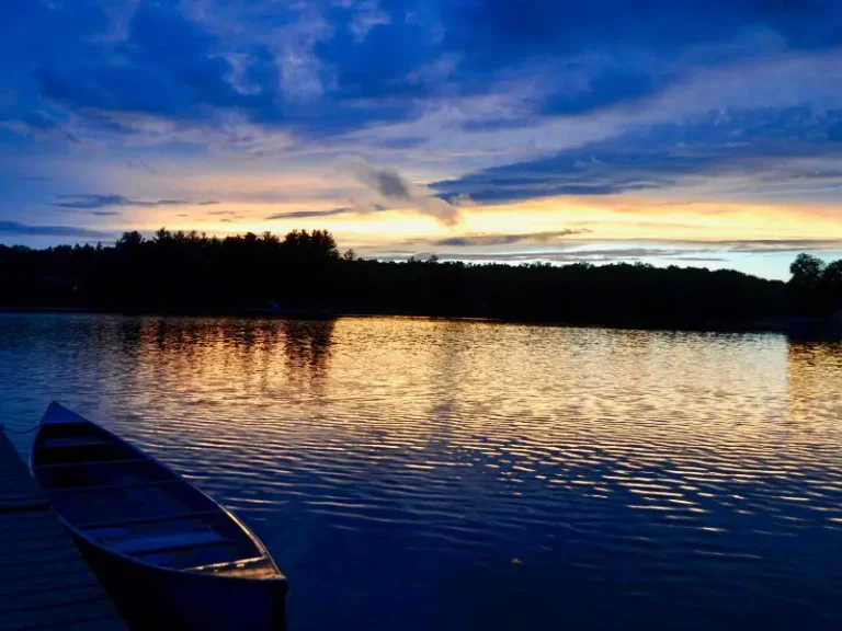 Canoe tied to a dock on a lake at sunset with trees silhouetted on the horizon.