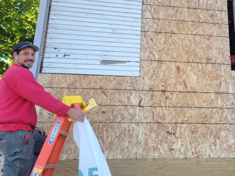 Man in a red sweatshirt holding a white bag next to an orange ladder and a plywood-covered wall.
