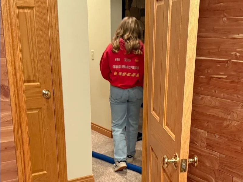 Person in a red hoodie and jeans walking away through a doorway in a wood-paneled room.