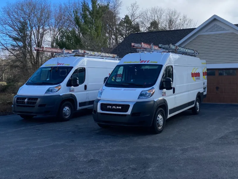 Two white work vans with ladders on top parked in a driveway near a house.
