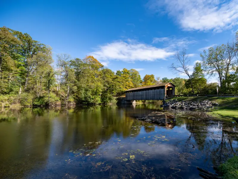 Covered wooden bridge over a calm river surrounded by trees under a blue sky.